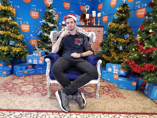 An image of Carl Reid in a chair wearing a Santa Hat, and Christmas Jumper. In the background there is a stack of Cool Blue boxes, and Christmas trees.
